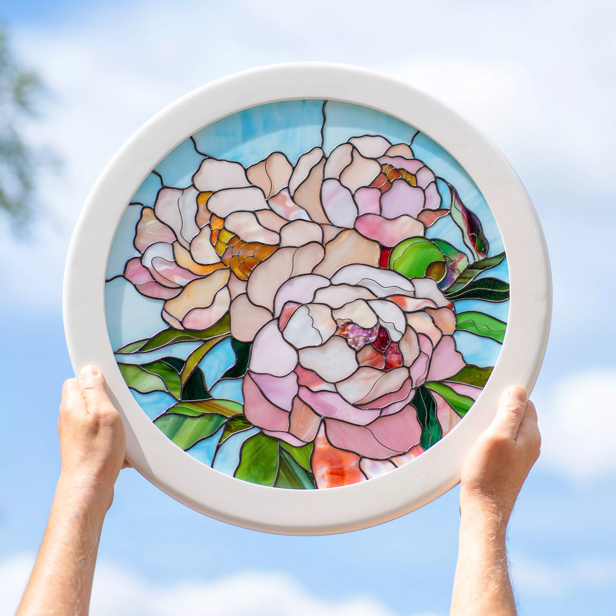 Handmade stained glass peony panel in a round white frame held up to a blue sky, blush-pink peonies with amber centers and green leaves glowing in the sunlight - floral glass wall art and gift