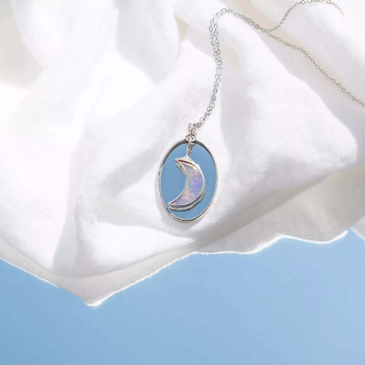 Handmade stained glass moon necklace on soft white fabric against a blue sky - an iridescent crescent moon glowing pink and lilac inside a round mirror pendant, on a fine stainless steel chain