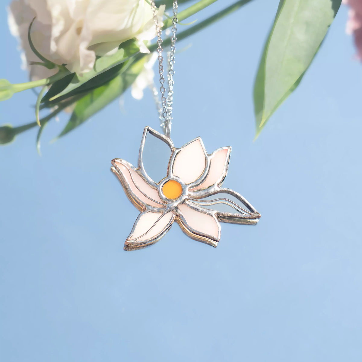 White Lotus stained glass necklace hanging in sunlight against a clear blue sky, the white and blush-pink glass petals and amber center glowing with soft cream flowers behind - handmade lotus pendant and a serene gift