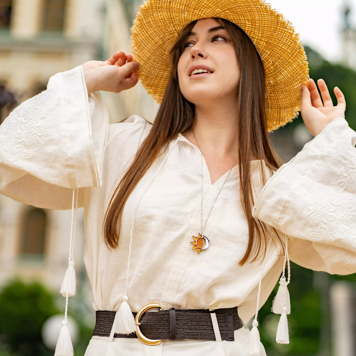 Smiling woman in a straw hat and white blouse outdoors wearing the handmade stained glass sun and moon necklace on a long chain - amber sun and silver crescent moon celestial jewelry for summer