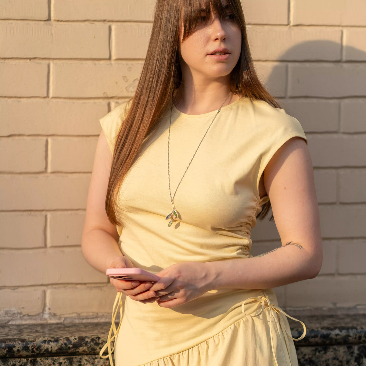 Woman in a yellow dress by a painted brick wall wearing the handmade stained glass leaf necklace long, the green glass branch pendant at her waist - botanical nature-inspired jewelry for everyday wear