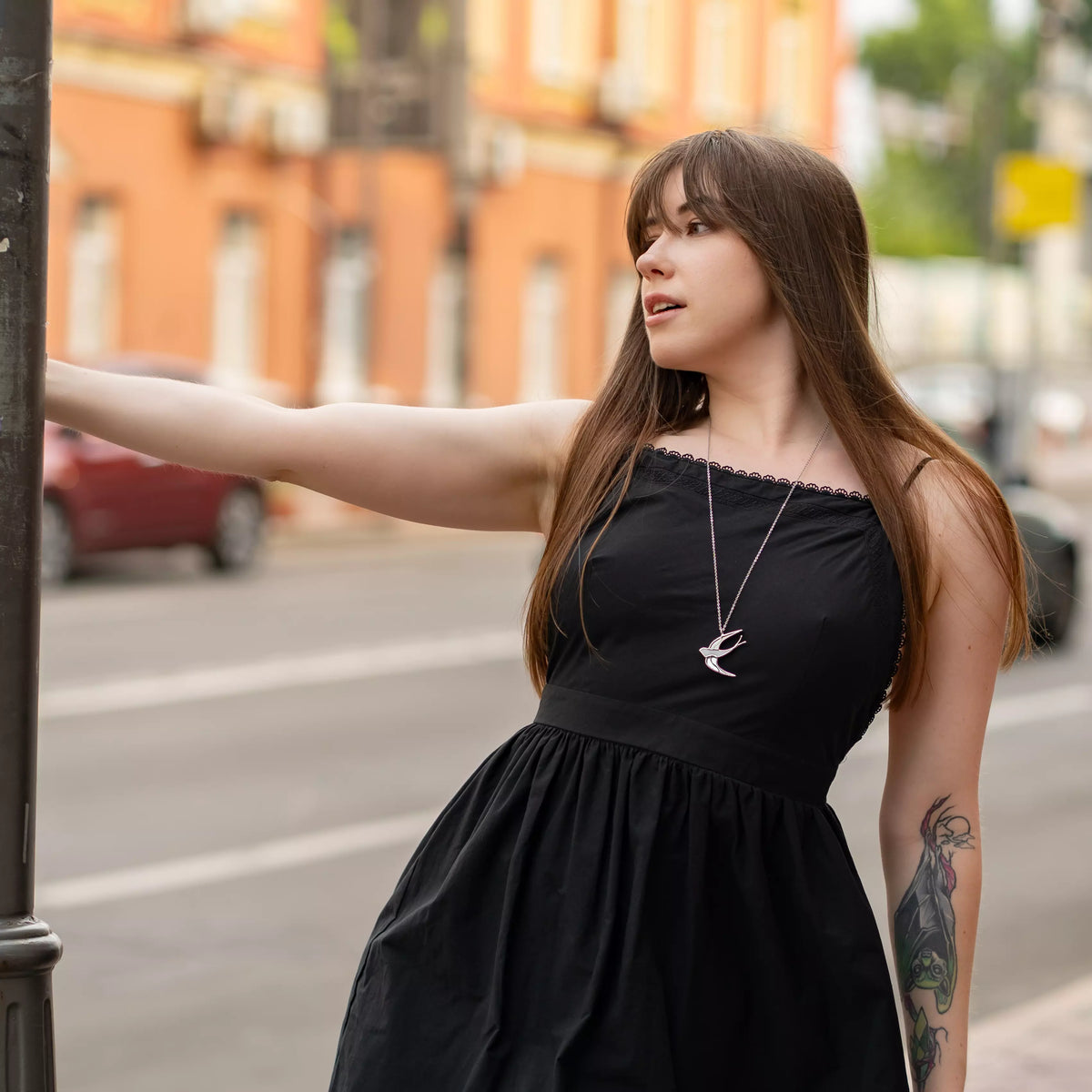 Model on a city street wearing the White Swallow stained glass necklace with a black dress - a long-chain handmade swallow pendant as everyday bird jewelry and a thoughtful gift