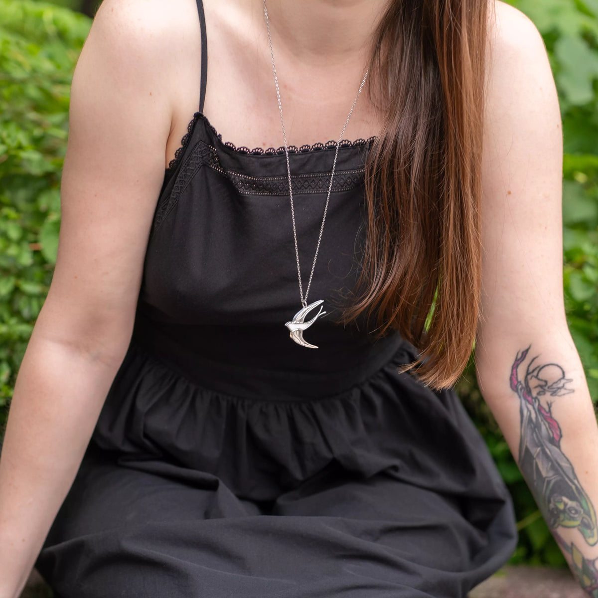 Seated woman wearing the White Swallow stained glass necklace on a long chain over a black dress with greenery behind - handmade stained glass swallow pendant and a meaningful gift