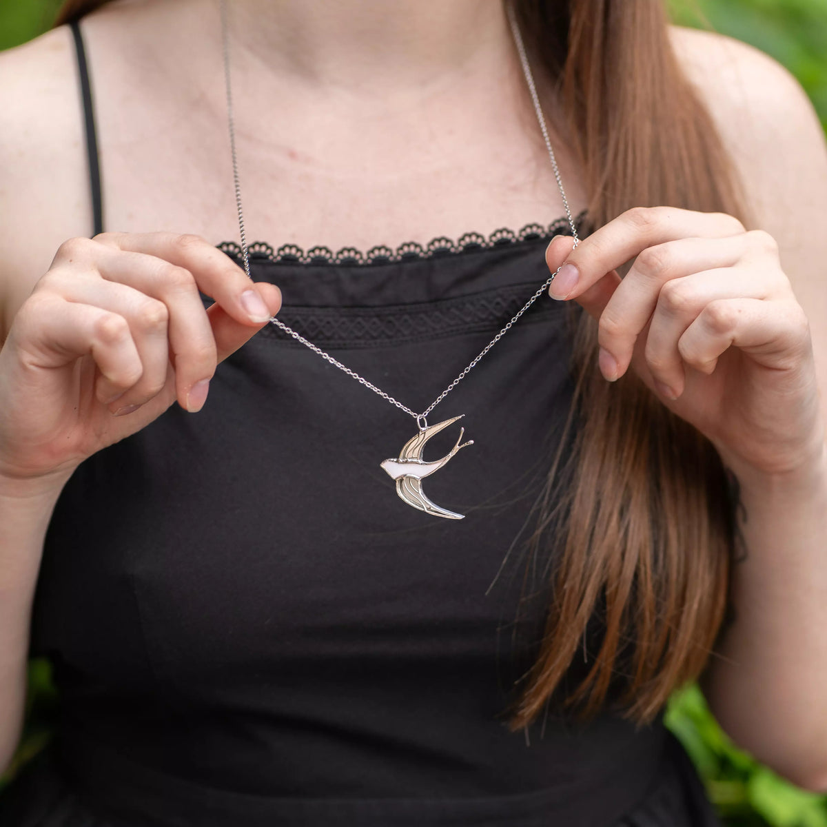 Close-up of a woman holding the chain of the White Swallow stained glass necklace, the white bird pendant resting on her black top outdoors - handmade stained glass bird jewelry and a gift