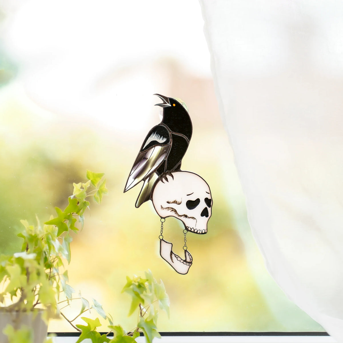 Raven and skull stained glass suncatcher on a sunlit window with a suction hook, ivy and a green garden beyond - cawing black raven on a bone-white skull with a hanging jaw, handmade gothic window decor