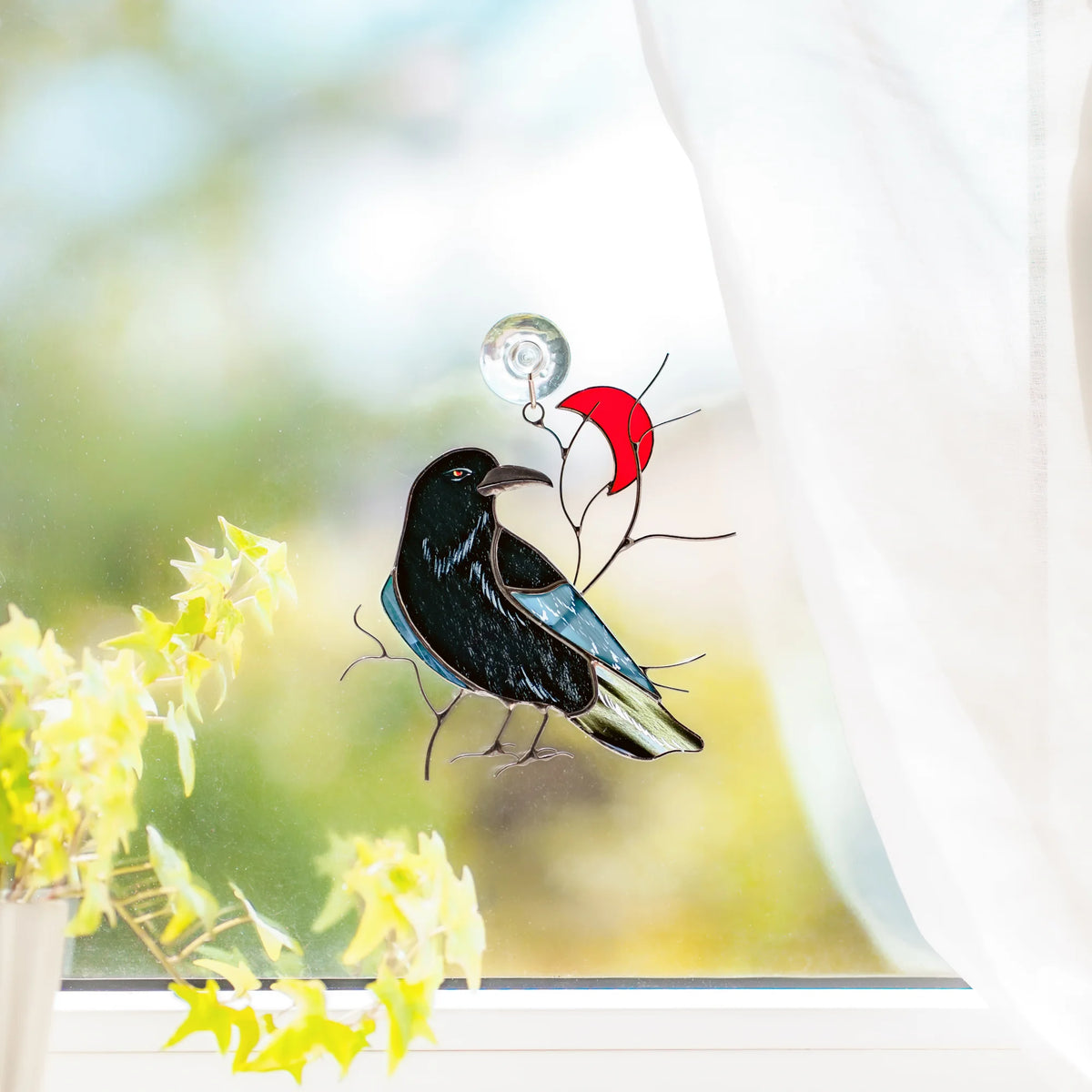 Raven and red crescent moon stained glass suncatcher on a bright window with a suction cup, sheer curtain and yellow-green foliage - handmade black raven with an iridescent teal wing, gothic window decor