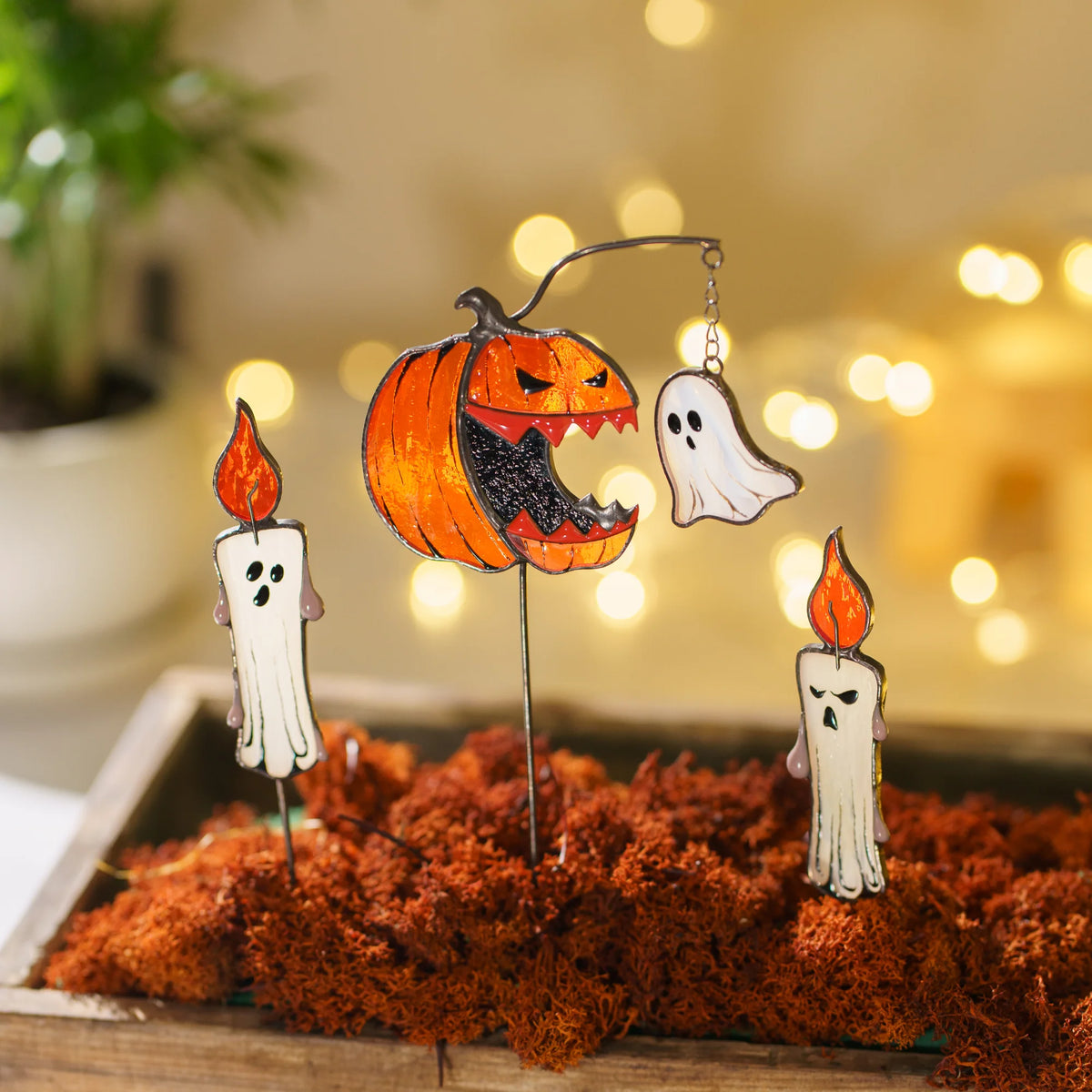 Halloween-themed stained glass plant stake decorations displayed in a tray of moss, featuring a carved jack-o'-lantern, two white candles with orange flames, and a white ghost, set against a warm blurred background with bokeh lights.