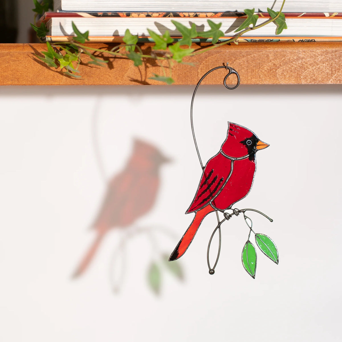 Red cardinal stained glass suncatcher hanging below a shelf of books and ivy, casting a soft cardinal shadow on the wall - handmade glass bird on a wire branch with green leaves