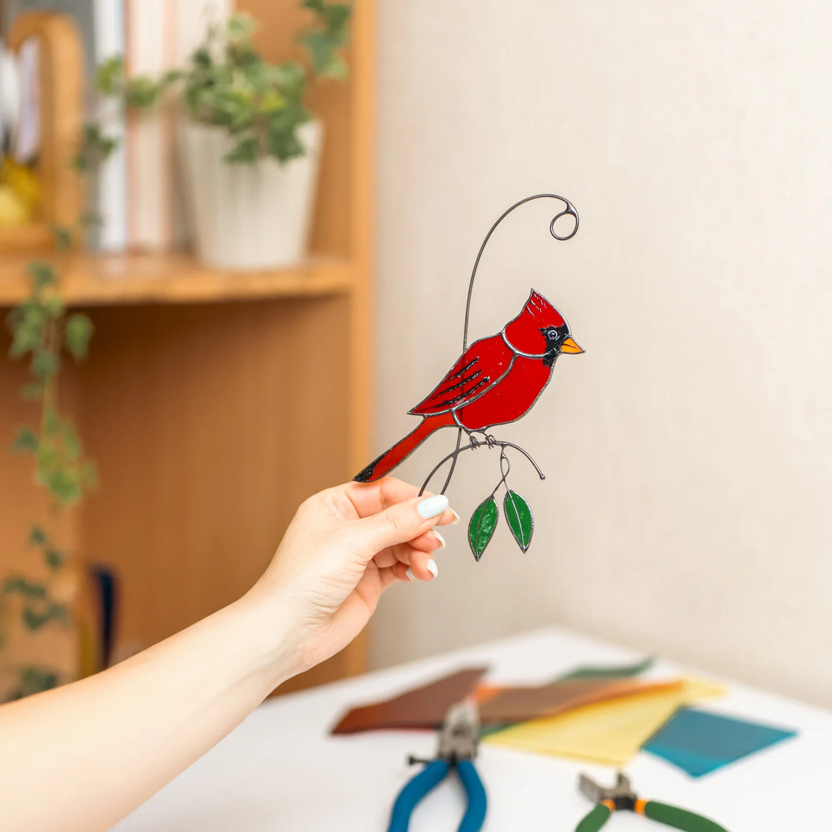 Hand holding a red cardinal stained glass suncatcher in a sunlit room, brilliant red cardinal on a wire branch with green glass leaves - handmade bird window decor and thoughtful gift