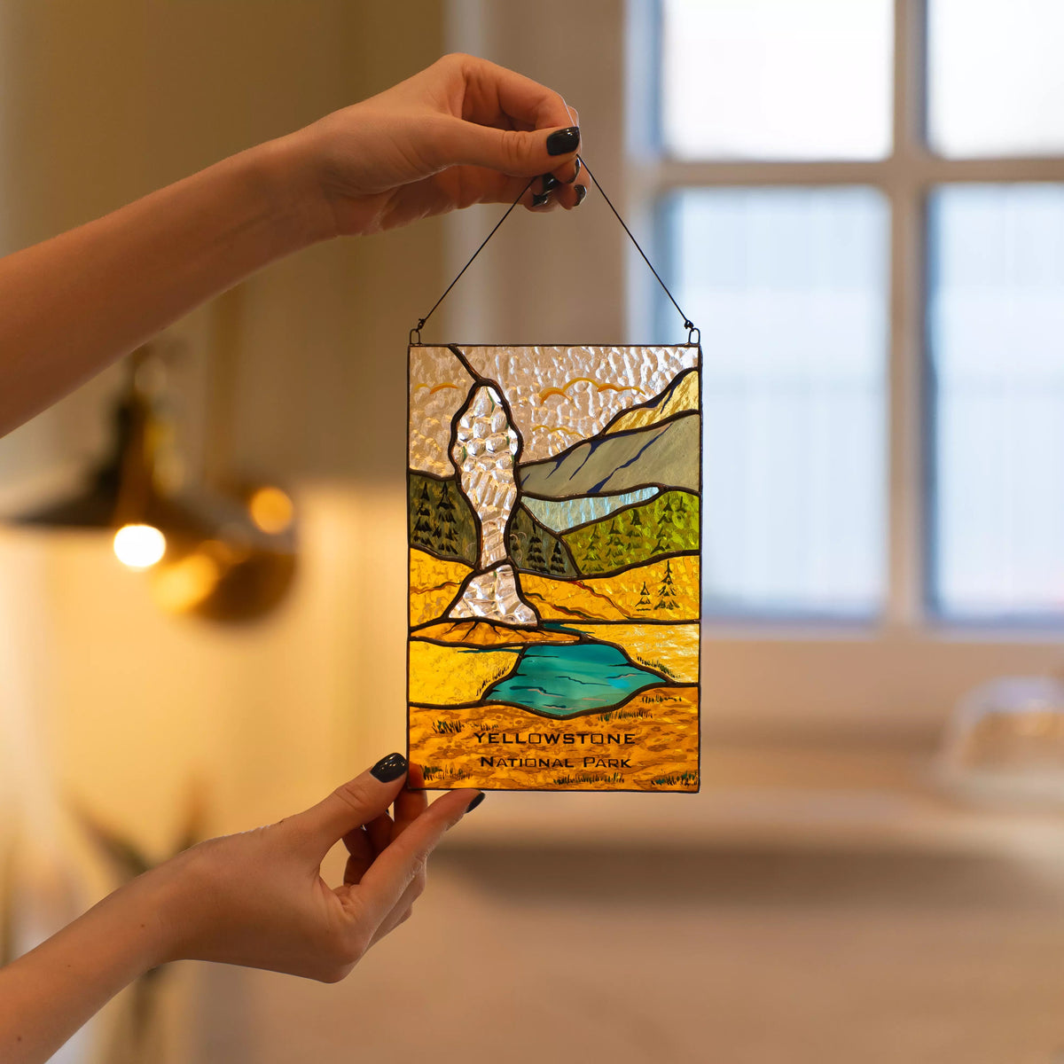 Hand holding a stained glass panel of Yellowstone National Park with a blurred indoor background