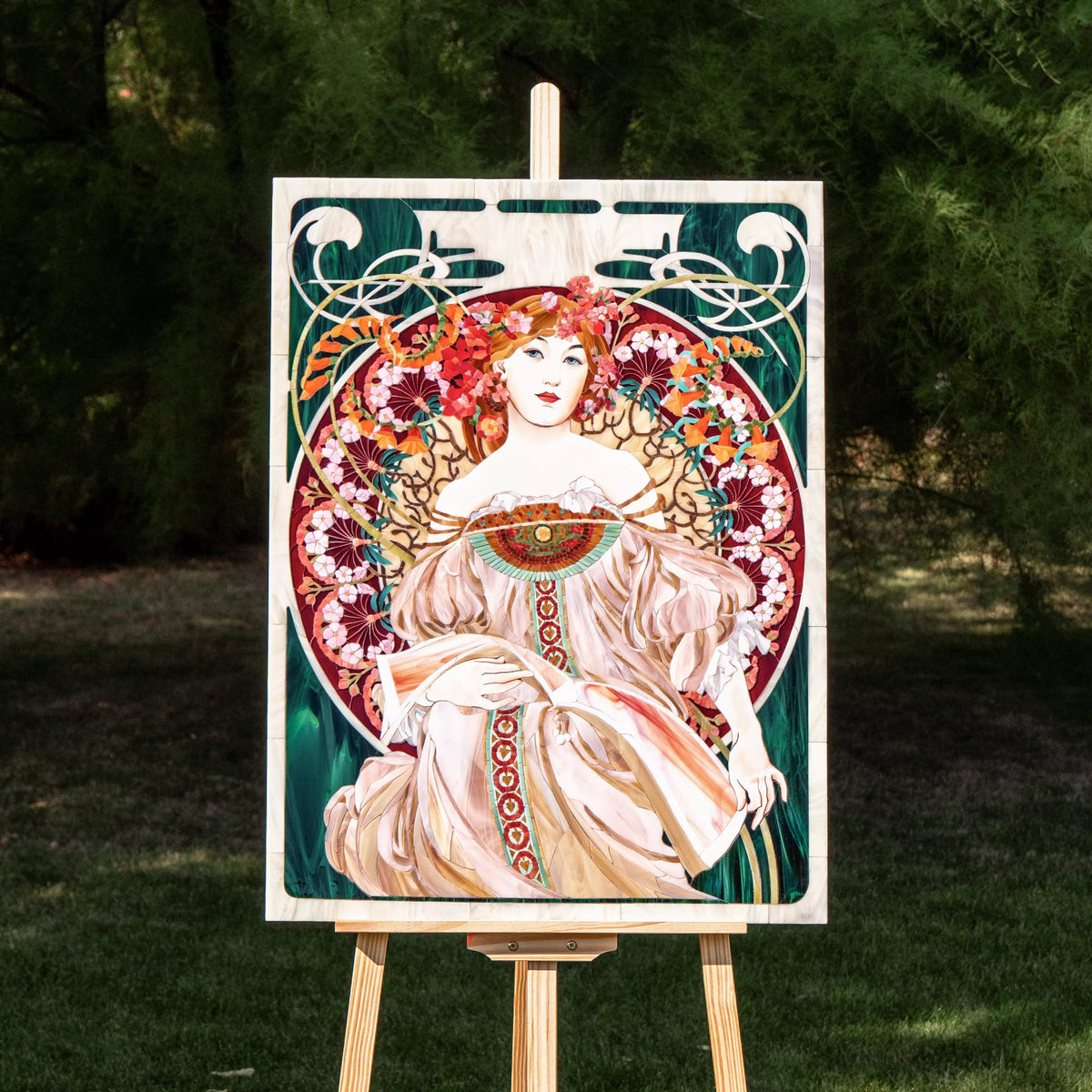Full view of Riviera mosaic artwork by Alphonse Mucha style, displayed outdoors on an easel, showing rich colors and detailed glass composition.