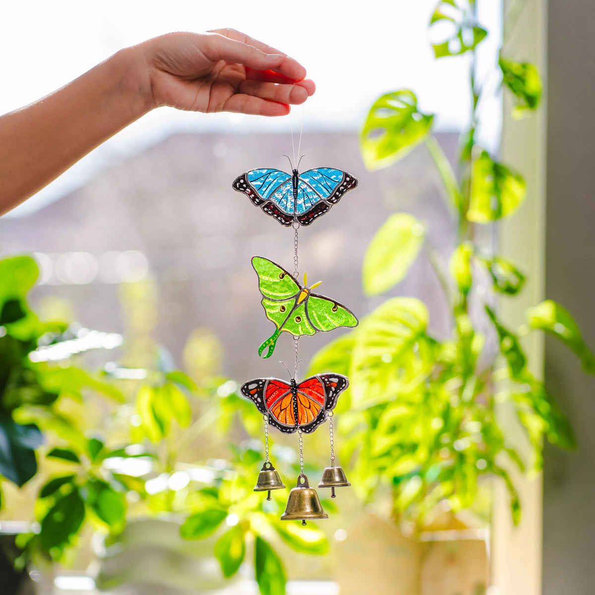 Butterfly stained glass wind chime with bells held by a hand against a blurred green plant background