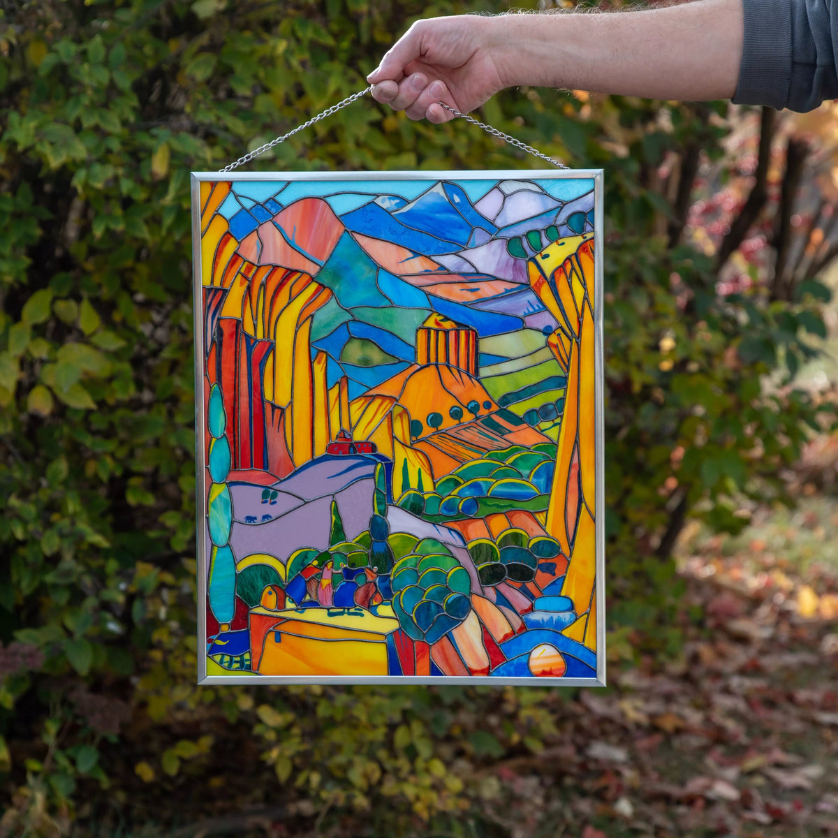 Colorful stained glass panel abstract landscape hanging outdoors with a person's hand holding it.