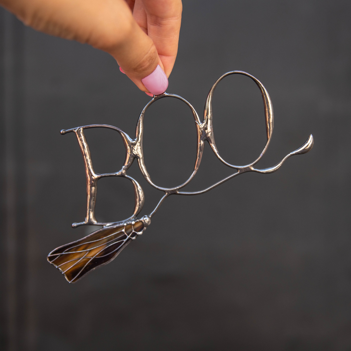 Close-up of a person holding a stained glass BOO suncatcher with orange broom against dark background