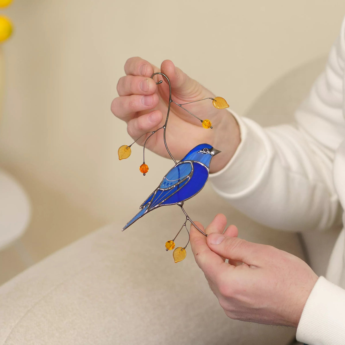 Hands holding stained glass indigo bunting bird ornament with yellow leaves on a beige background