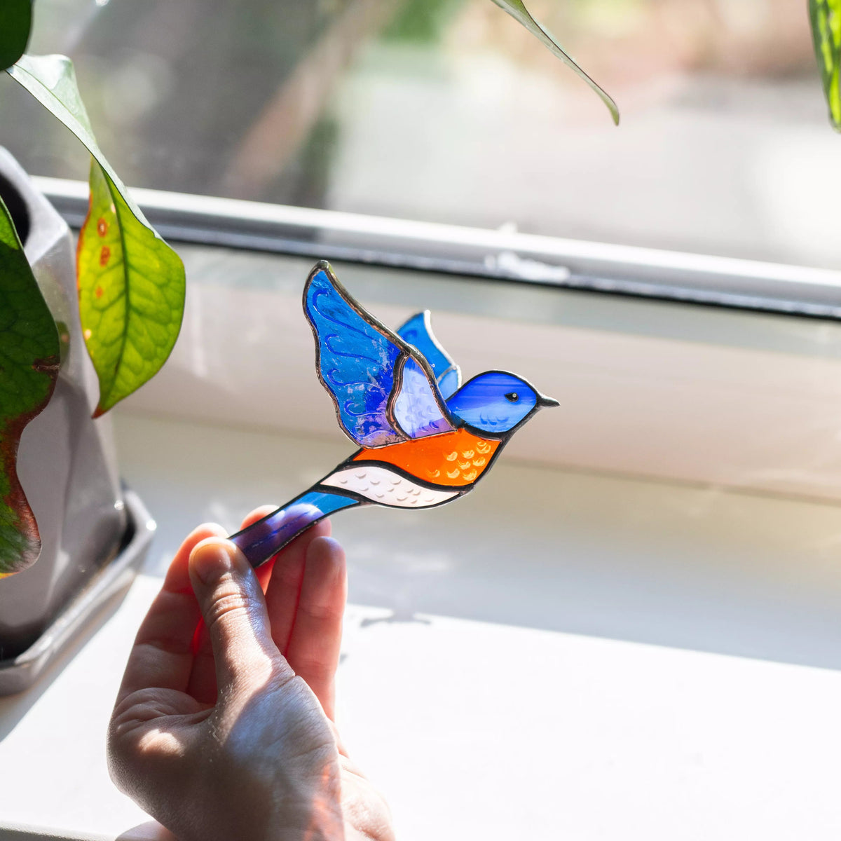 Hand holding a colorful stained glass bluebird by a window