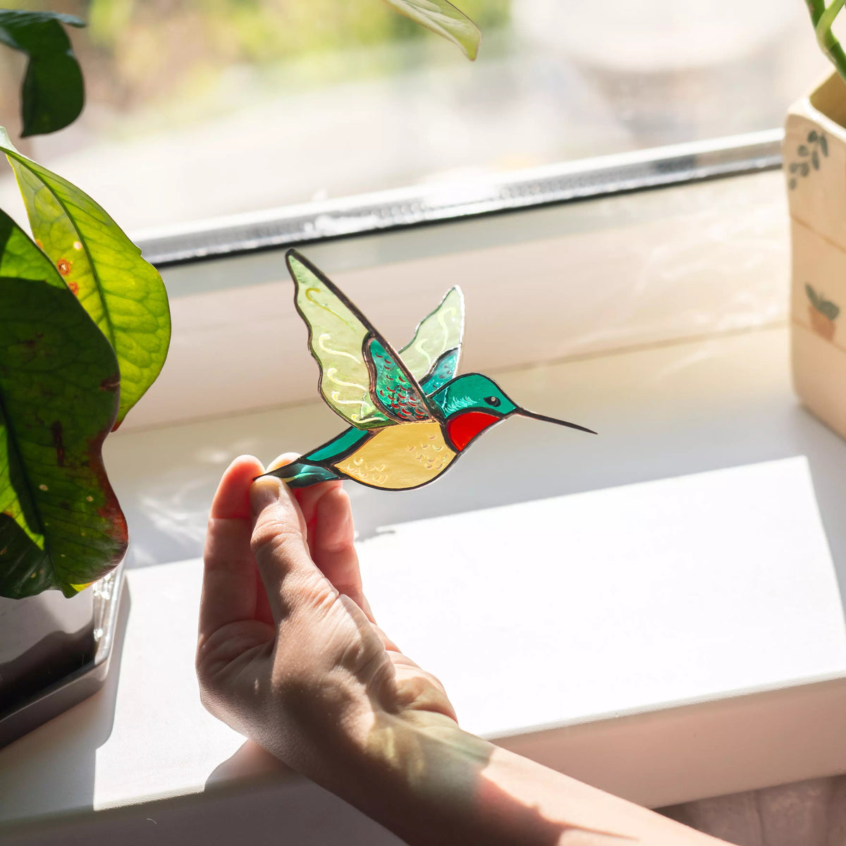 Hand holding a colorful stained glass hummingbird suncatcher by a window