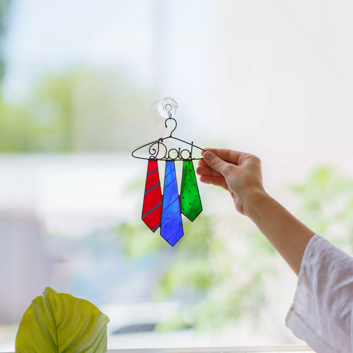 Hand holding a stained glass ornament with red, blue, and green tie designs in front of a window.