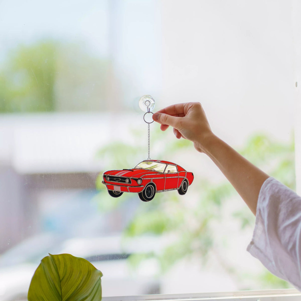 Hand holding a red car stained glass suncatcher in front of a window with a blurred outdoor view.