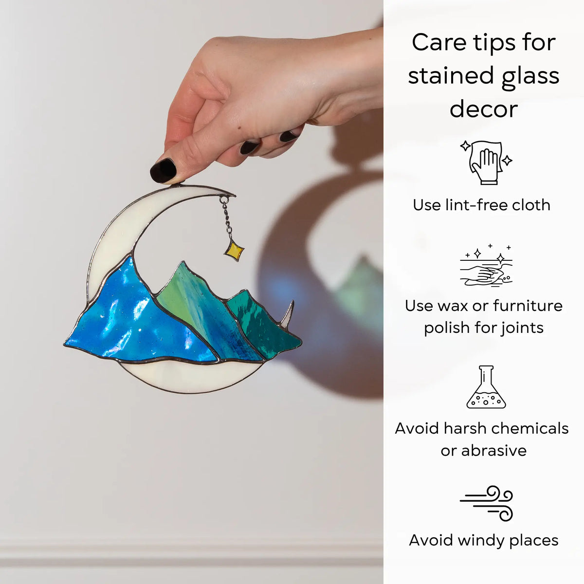 Hand holding stained glass celestial suncatcher moon and blue mountains with care tips beside: use lint-free cloth, polish joints with car wax or furniture polish, avoid harsh chemicals, and windy places.