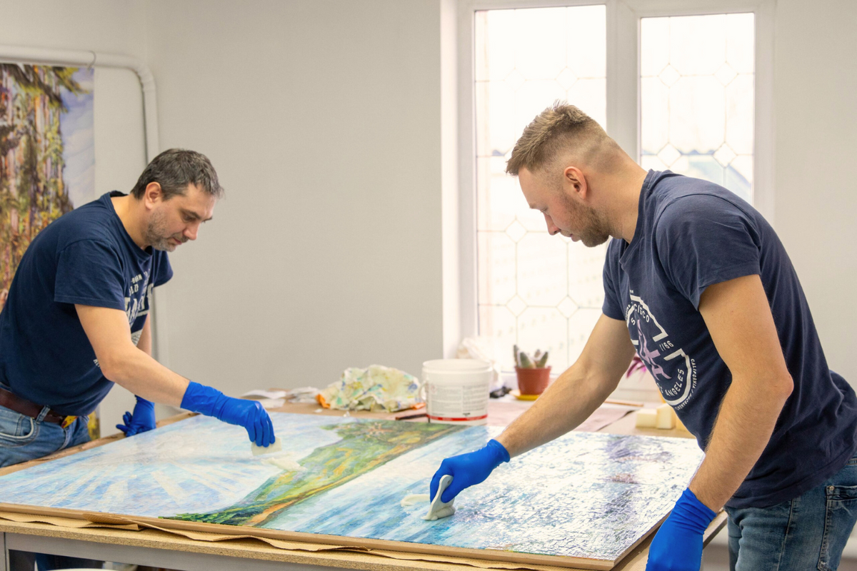 Two men wearing blue gloves work on a large painting in a studio