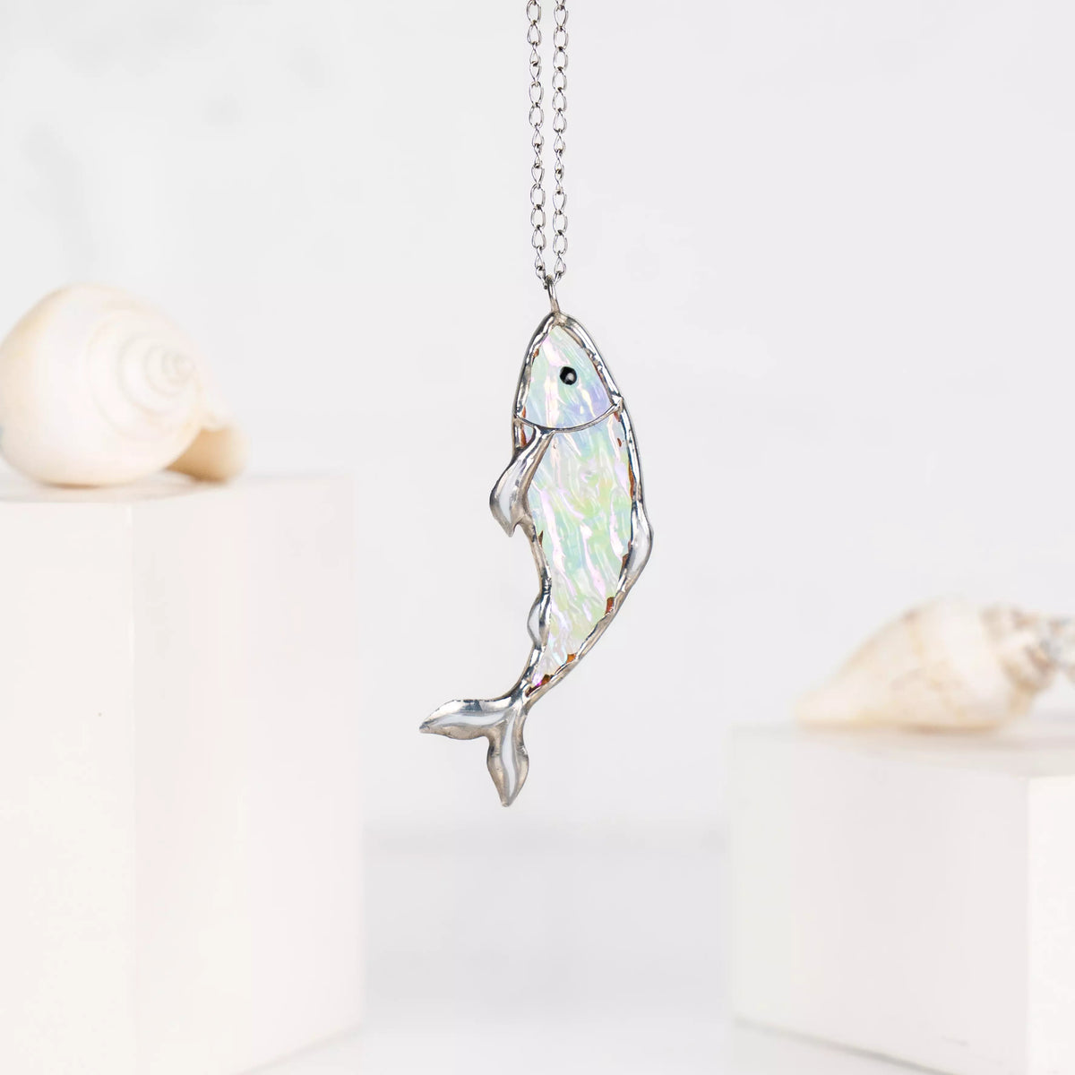 Clear stained glass fish necklace in light tones, shown with soft focus seashells in the background