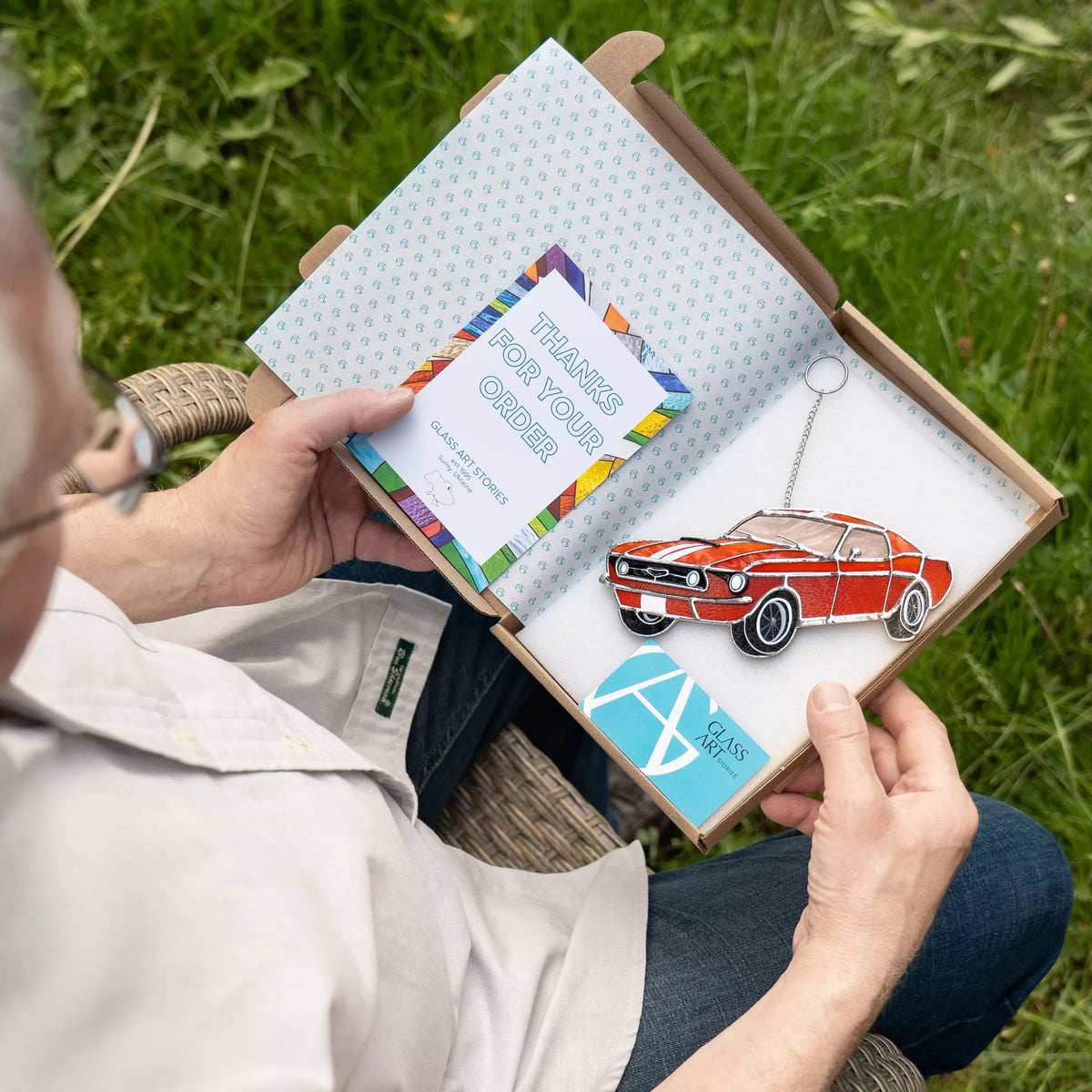 Person holding a box with a car stained glass suncatcher and a thank you card outdoors.