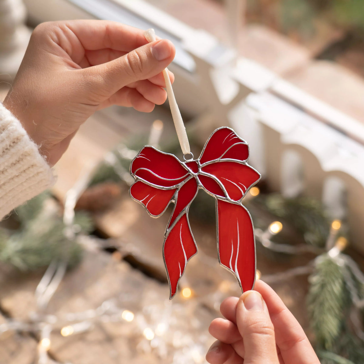 Red bow stained glass ornament held by a person with blurred Christmas lights and decorations in the background