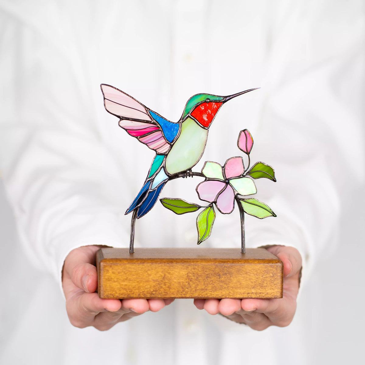 Hands holding stained glass hummingbird sculpture against a white background