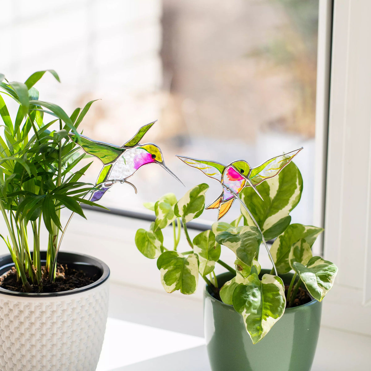 Two potted plants with stained glass hummingbird plant stake on a windowsill.
