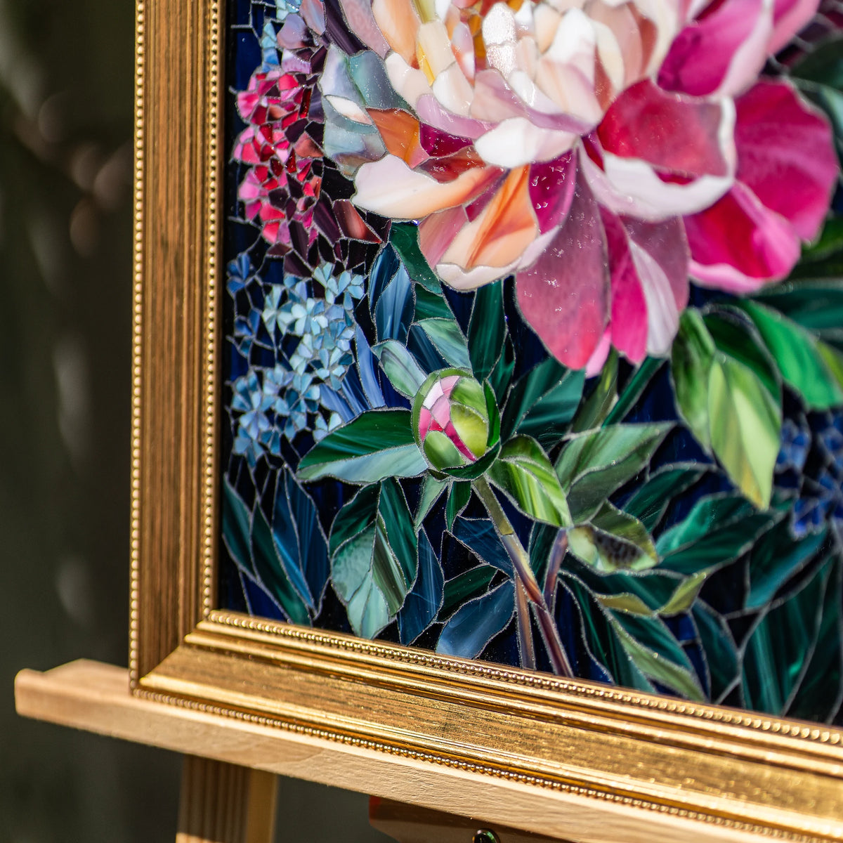 Close-up of a peony stained glass mosaic showing hand-cut glass petals, blue hydrangea and a peony bud against navy glass in a beaded gold frame — detail of handmade floral wall art