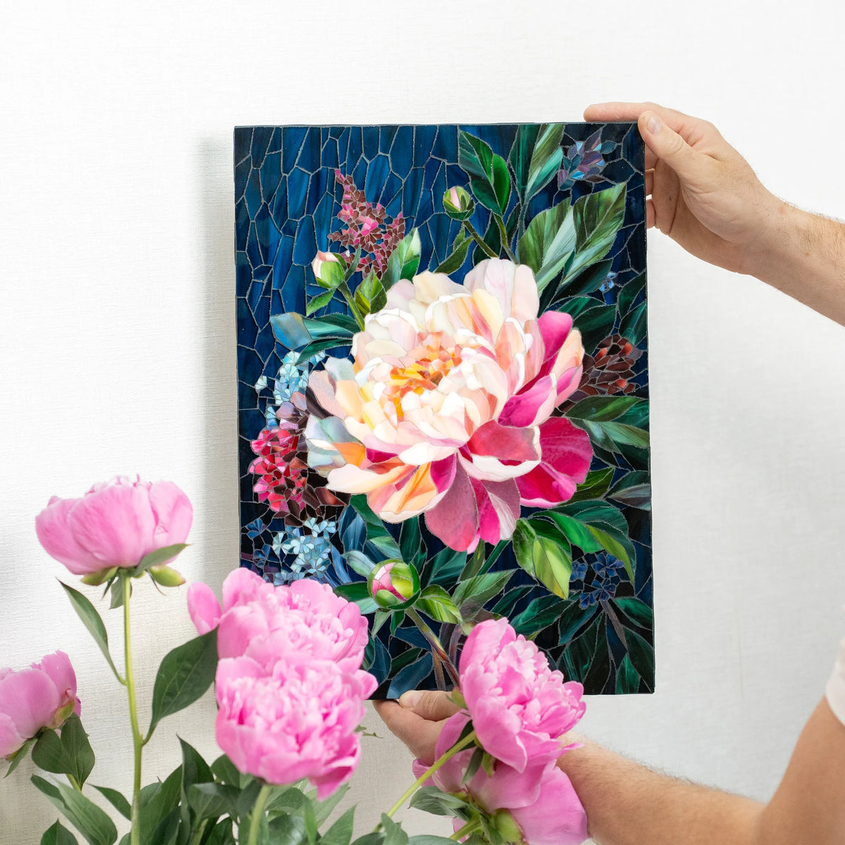 Hands holding a peony stained glass mosaic beside fresh pink peonies, showing how lifelike the hand-cut glass petals look — handmade stained glass floral wall art for the home