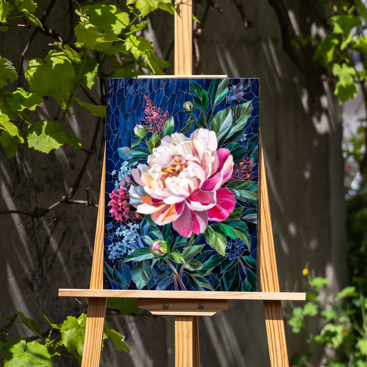 Peony stained glass mosaic panel on a wooden easel among grapevines outdoors, pink bloom and green foliage lit by sun — handmade stained glass floral wall art and a unique gift
