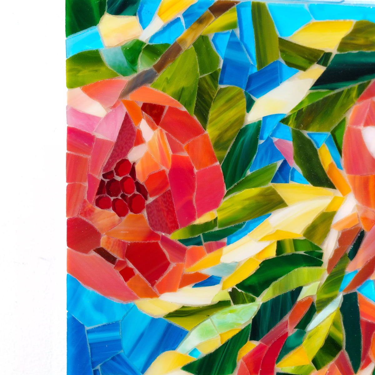 Close-up of the pomegranate stained glass mosaic showing a split fruit with ruby-red glass seeds, hand-cut red and coral tiles, green leaves and grout lines against blue glass - detail of handmade glass wall art