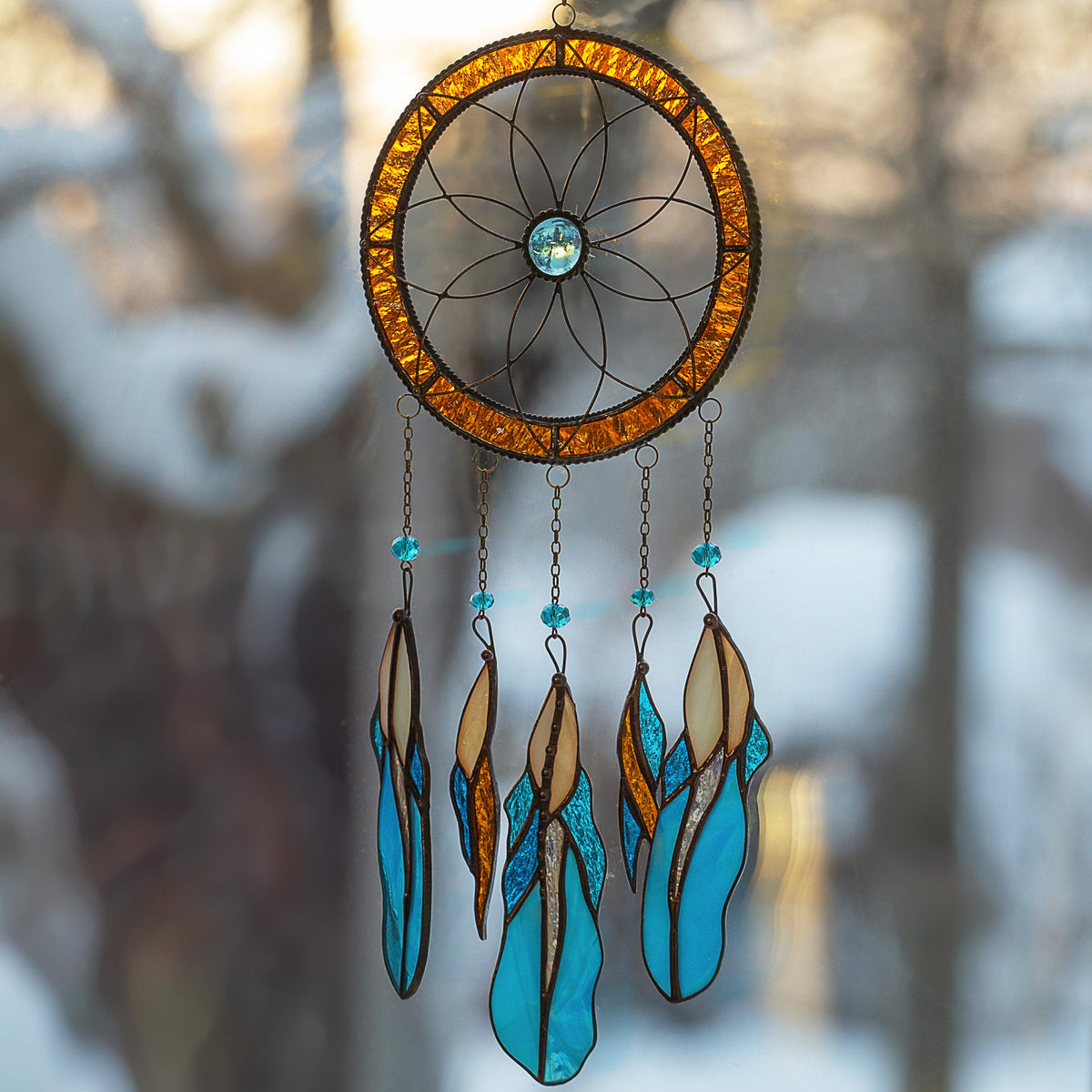 Dreamcatcher glowing in warm light, hanging in a softly lit background with snow-covered trees visible through the window.